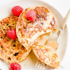 2 pieces of French toast on white plate with syrup and raspberries and a small piece on a fork