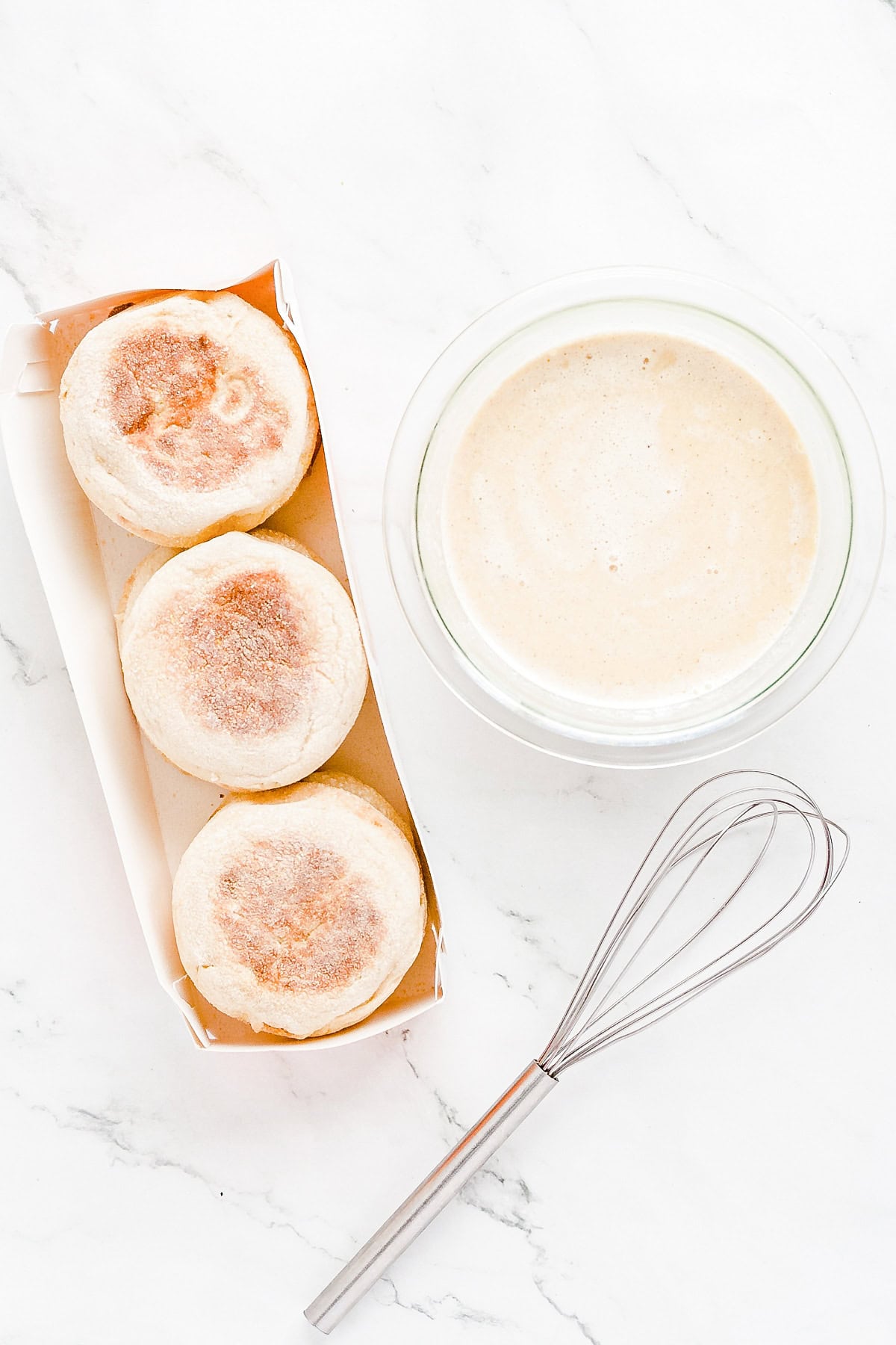 Pack of English muffins alongside bowl of custard and a wire whisk