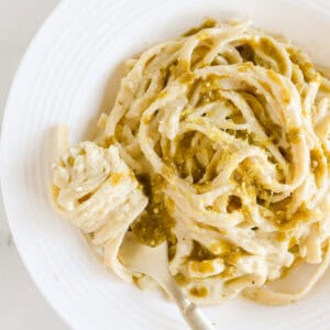 White plate of pesto alfredo pasta garnished with extra pesto and lemon zest.