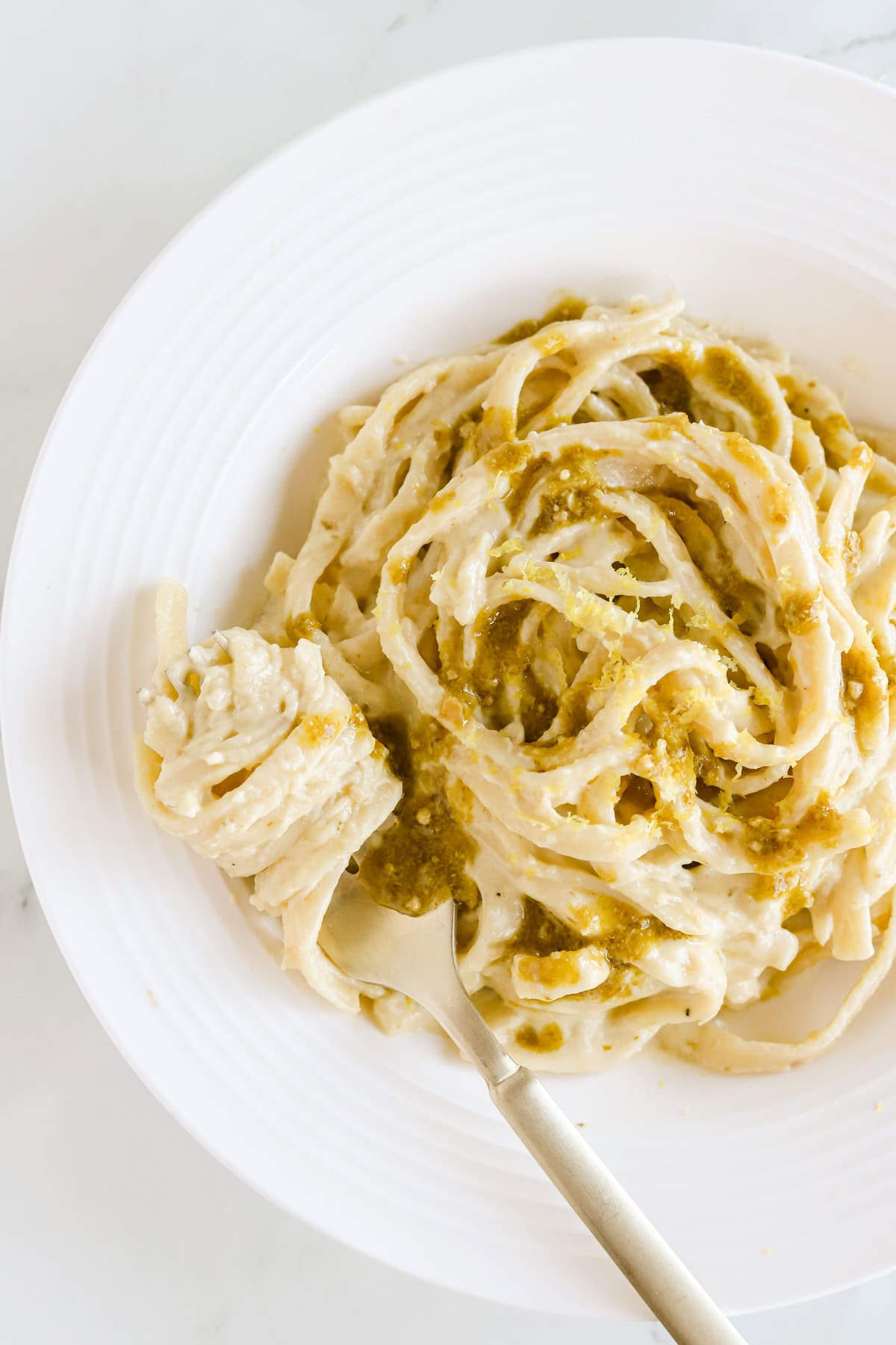 White plate of pesto alfredo pasta garnished with extra pesto and lemon zest.