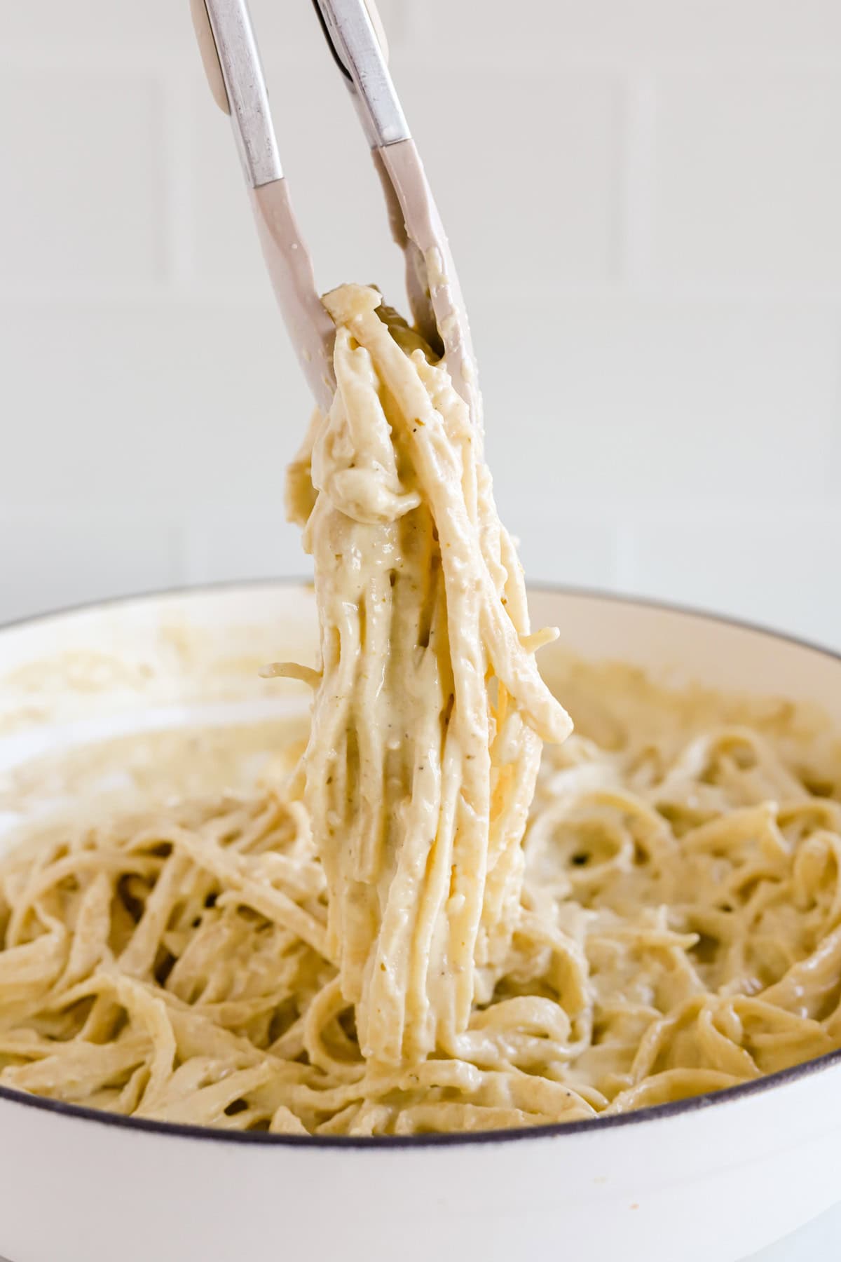Tongs lifting fettuccine noodles coated with pesto alfredo sauce out of skillet.