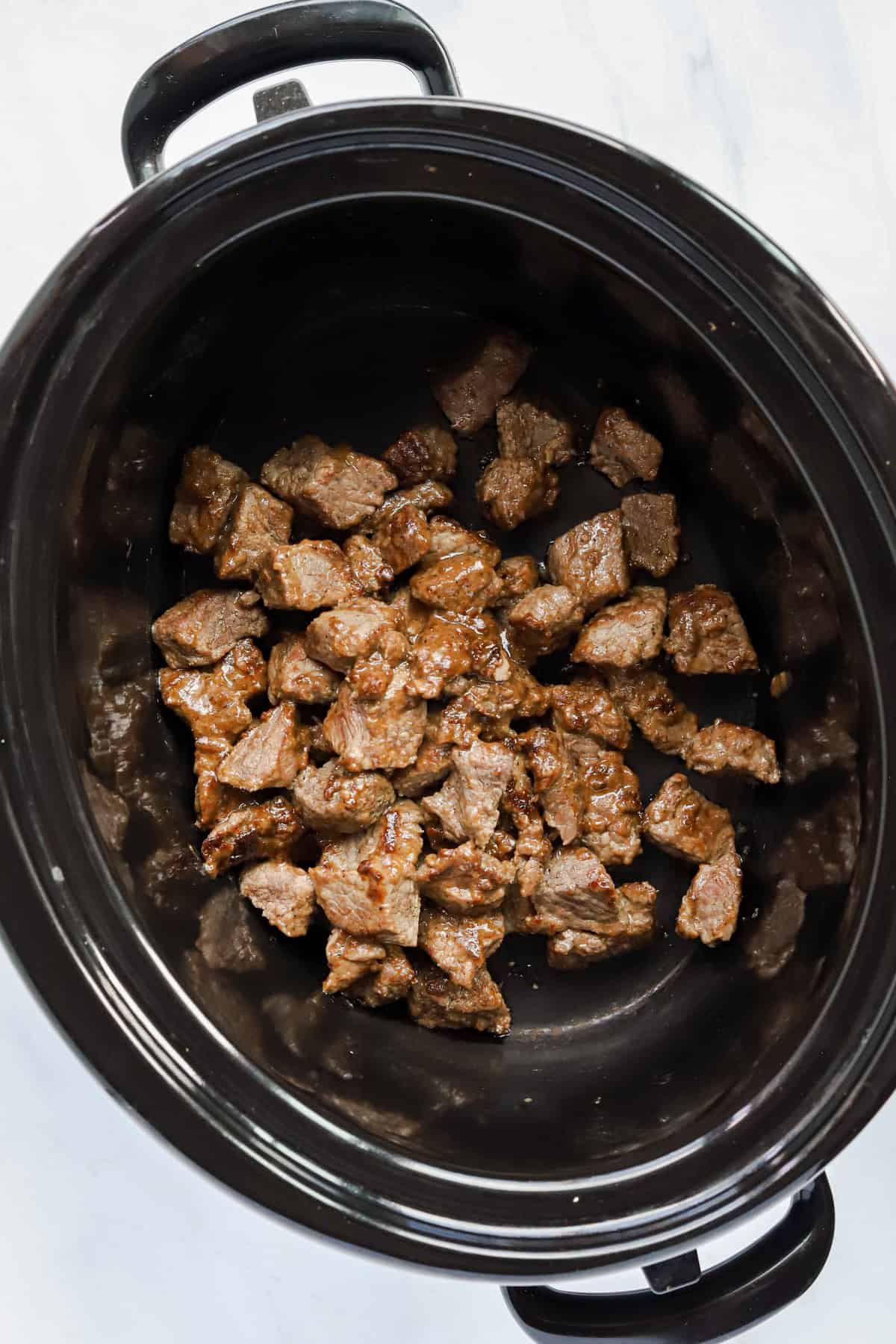 Browned chuck roast chunks in the bottom of a slow cooker.