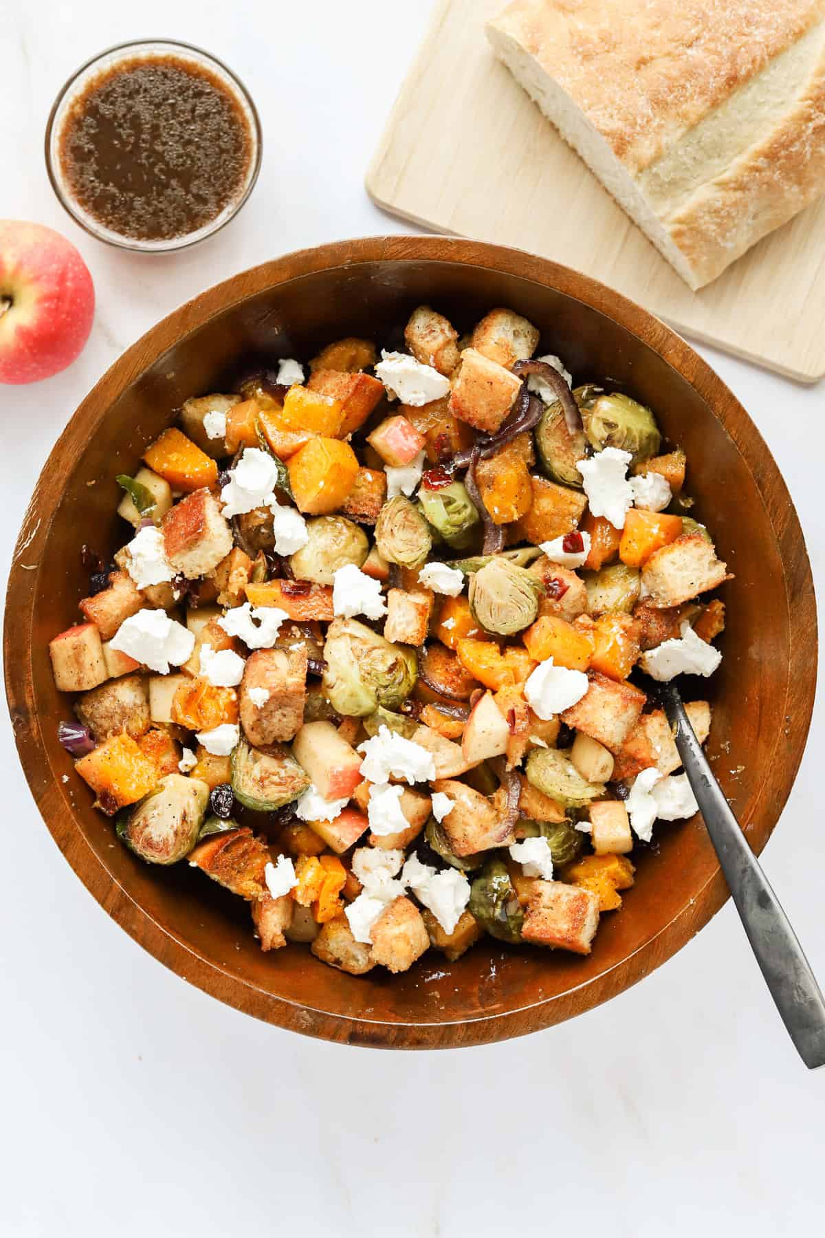 Wooden bowl of fall panzanella salad with a black spoon surrounded by italian loaf on a wooden board, extra vinaigrette cup and an apple.