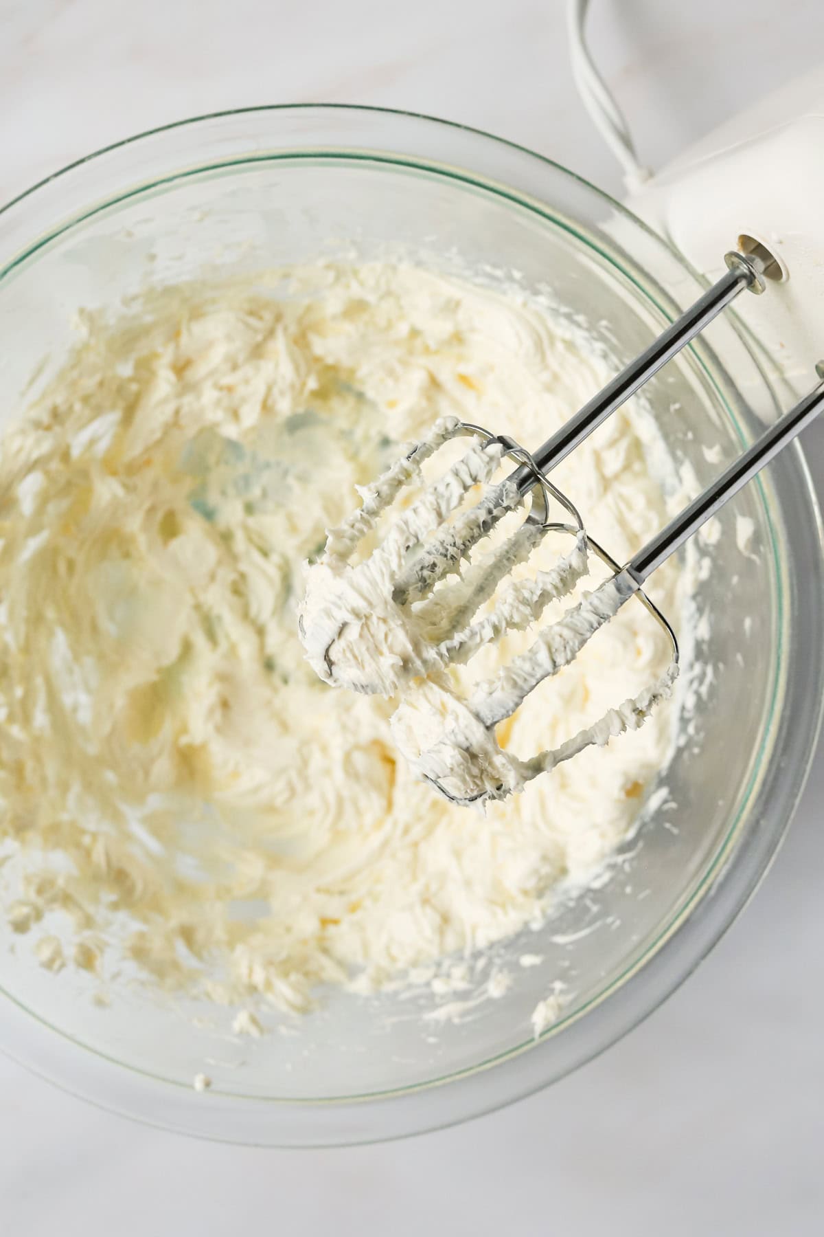 Hand mixer resting on countertop covered in cream cheese over bowl.