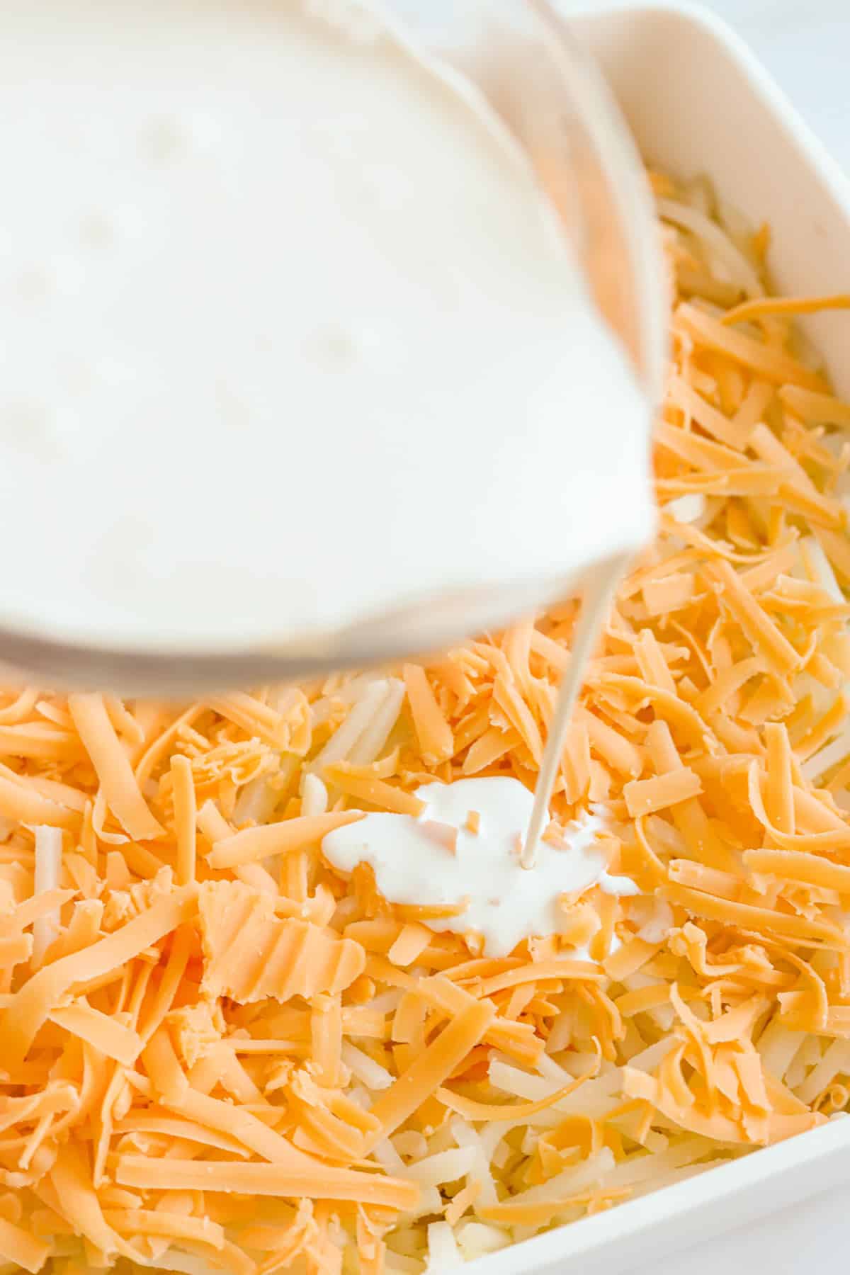 Cream being poured from glass measuring cup over cheese and hash browns in white baking dish.