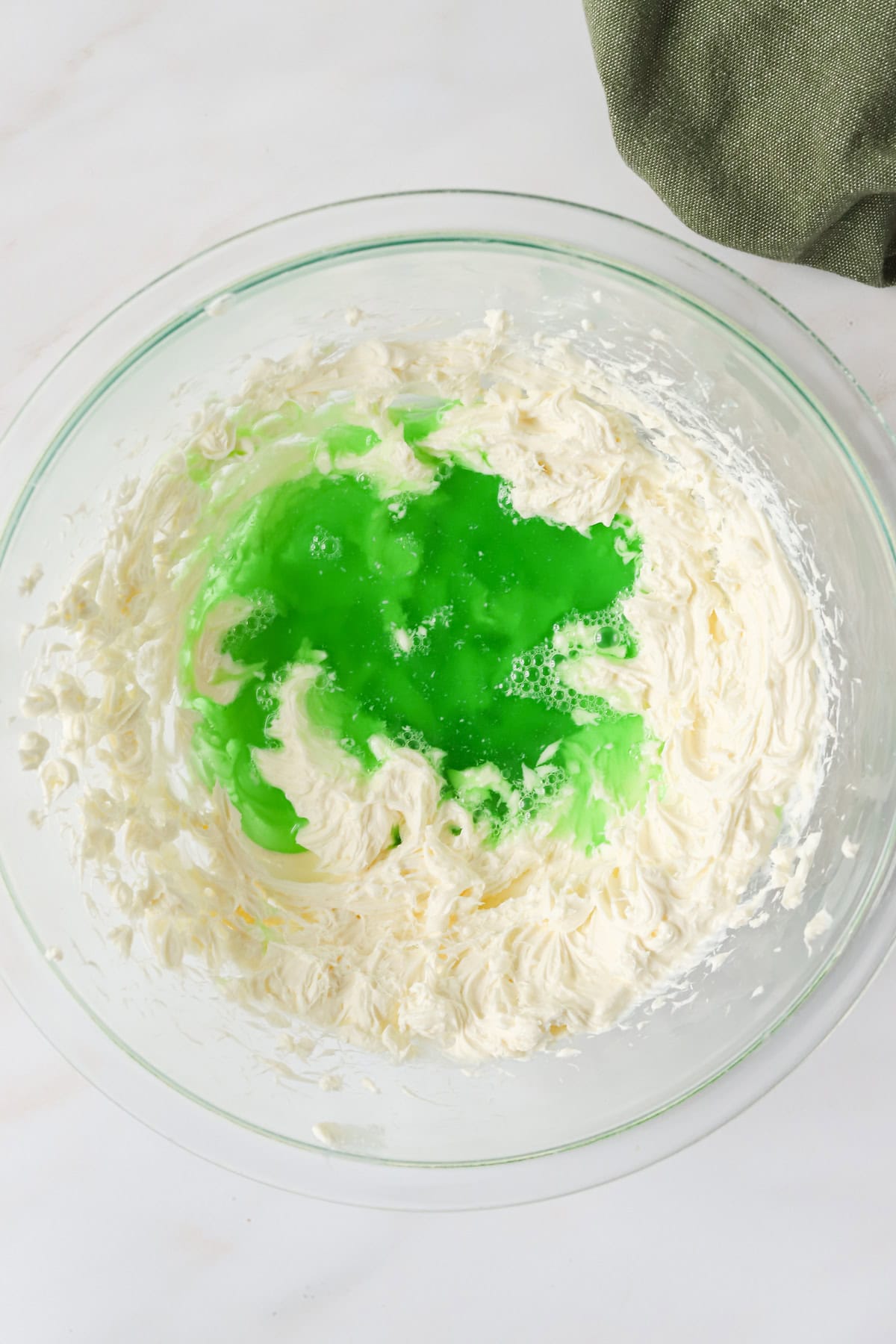 Hot lime jello mixture added to whipped cream cheese in a glass bowl.