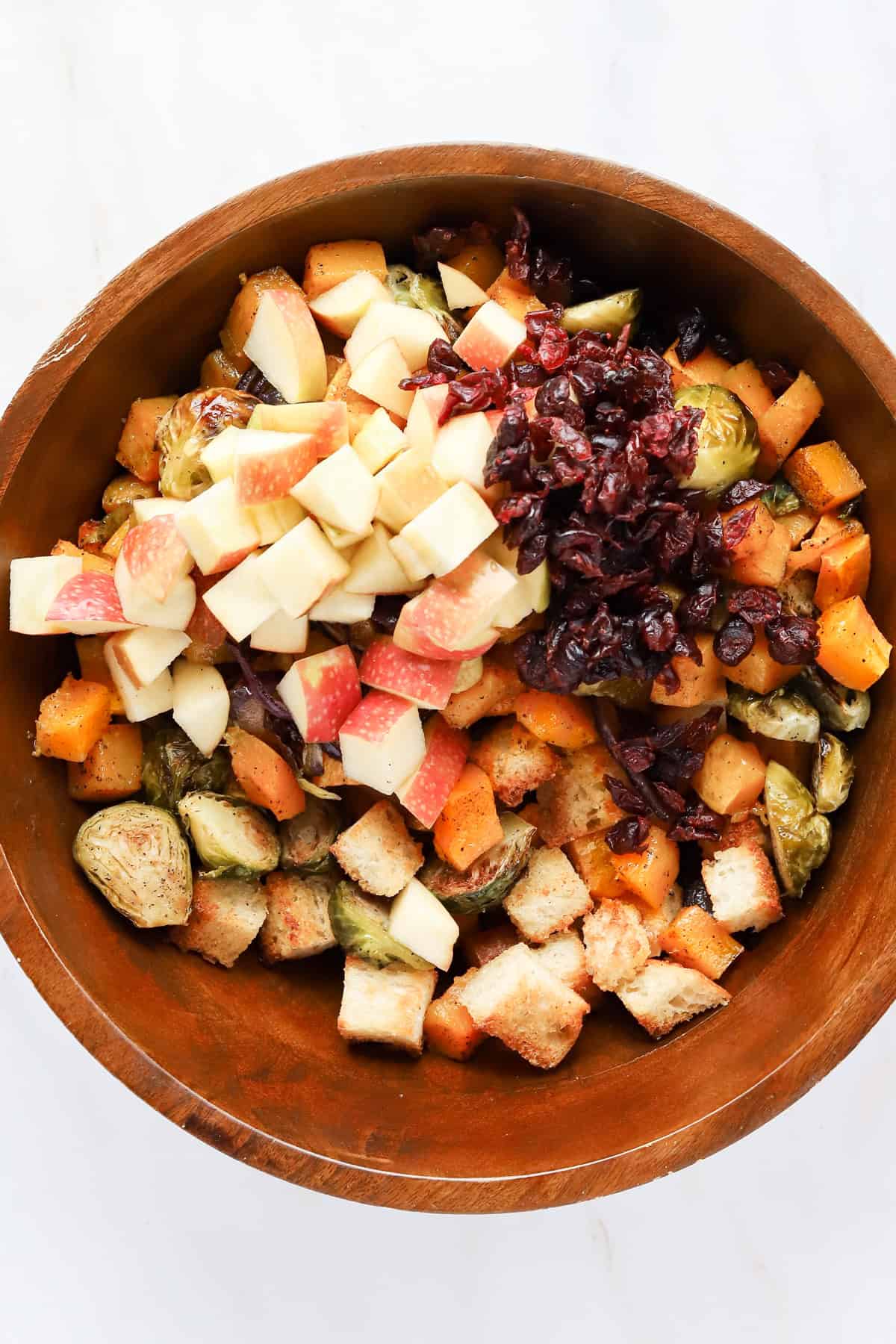 Overhead view of croutons, roasted vegetables, dried cranberries and diced apples in wooden bowl.