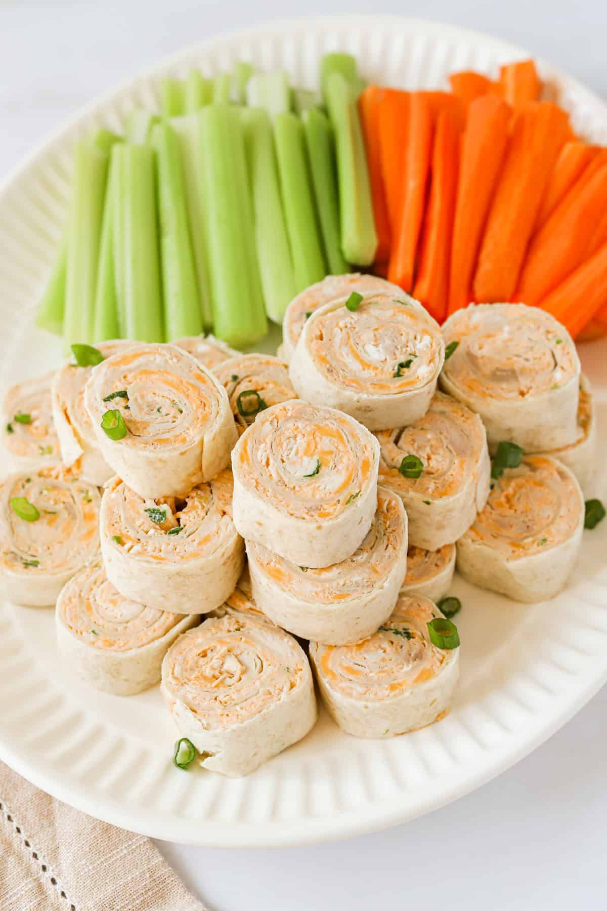 Buffalo chicken pinwheels piled neatly on a white platter with carrot and celery sticks behind them