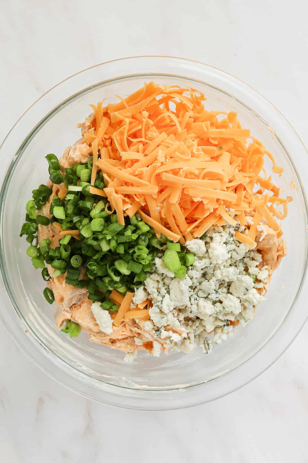 Shredded cheddar, blue cheese crumbles and sliced green onions in bowl with buffalo chicken mixture.