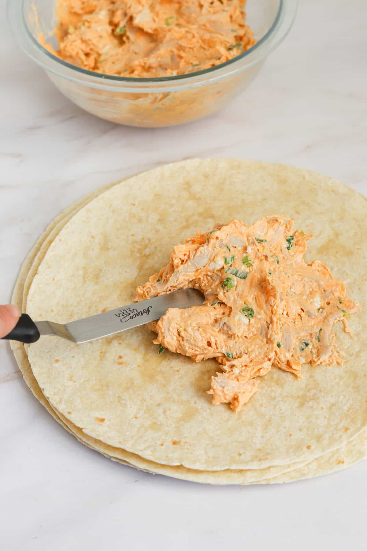 Metal spatula spreading filling over tortilla.