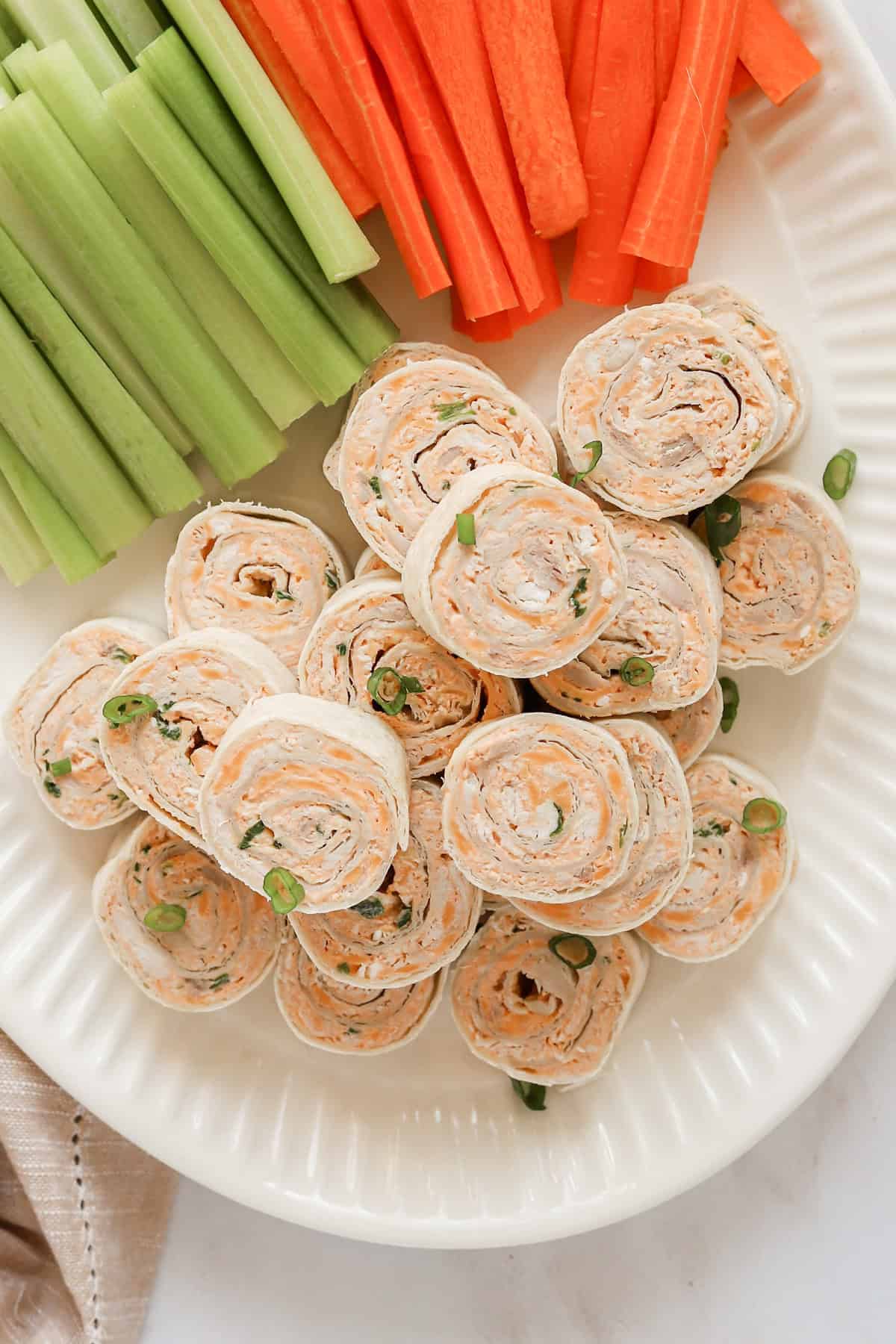 Overhead shot of buffalo chicken pinwheels piled on a white platter with carrot and celery sticks