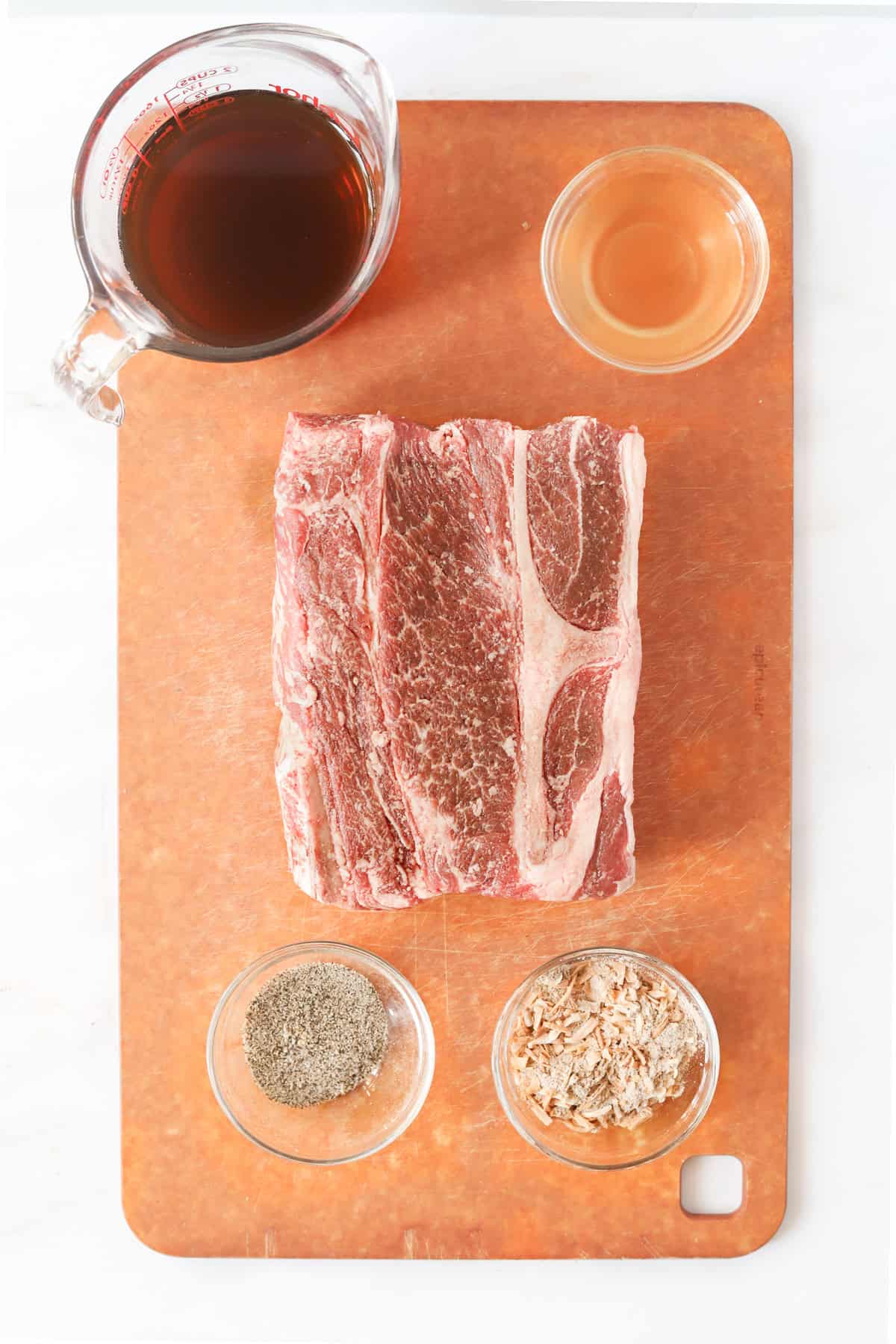 Chuck roast on wooden cutting board with beef broth, apple cider vinegar, spices and onion soup mix in bowls surrounding it.