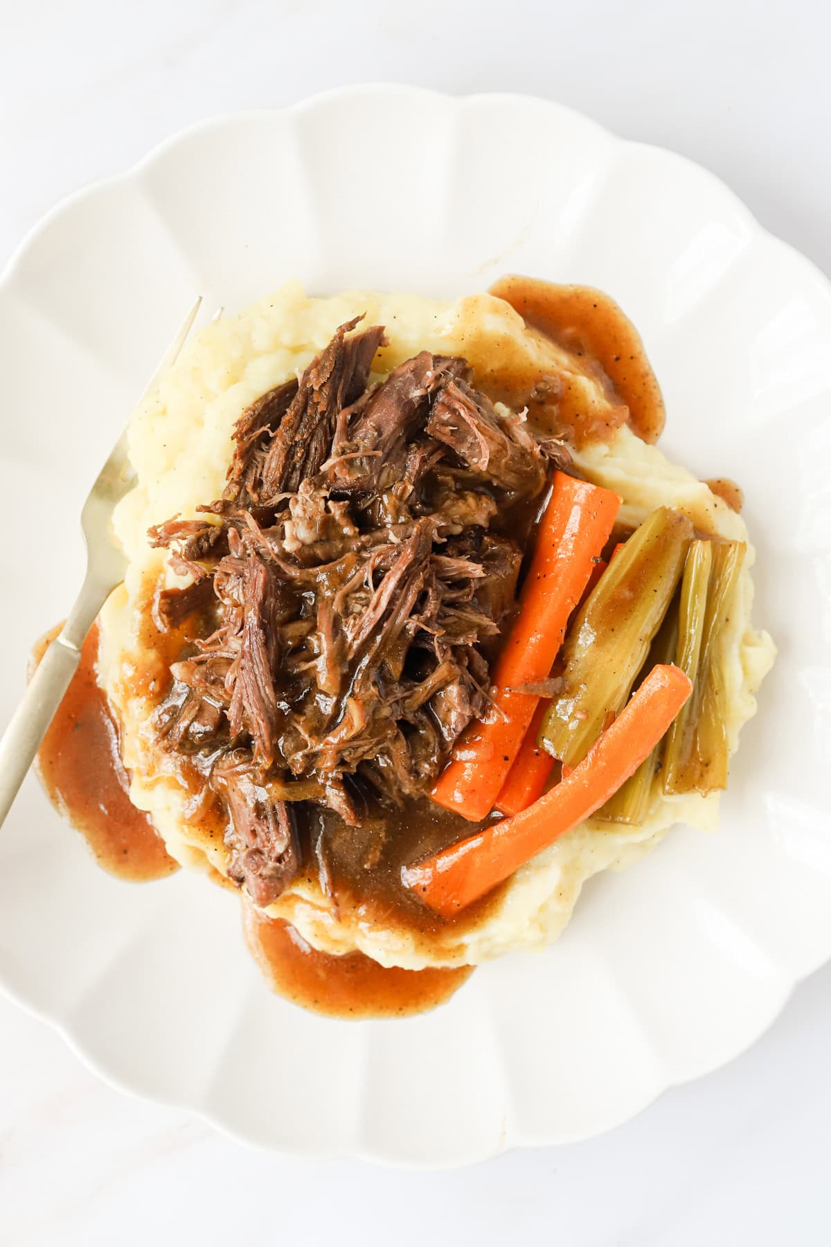 Plated pot roast, mashed potatoes, carrots and celery on a scalloped white plate.
